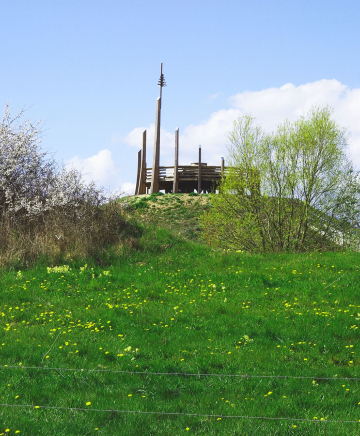 Grüner Hügel mit einer Plattform aus Holz an der Spitze, im Hintergrund blühende Bäume im Frühling