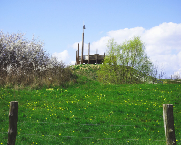 Grüner Hügel mit einer Plattform aus Holz an der Spitze, im Hintergrund blühende Bäume im Frühling