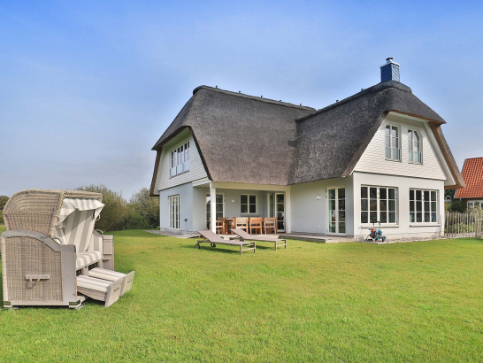 Reetgedecktes Ferienhaus mit Terrasse in einem  Garten und einem Strandkorb im Vordergrund.